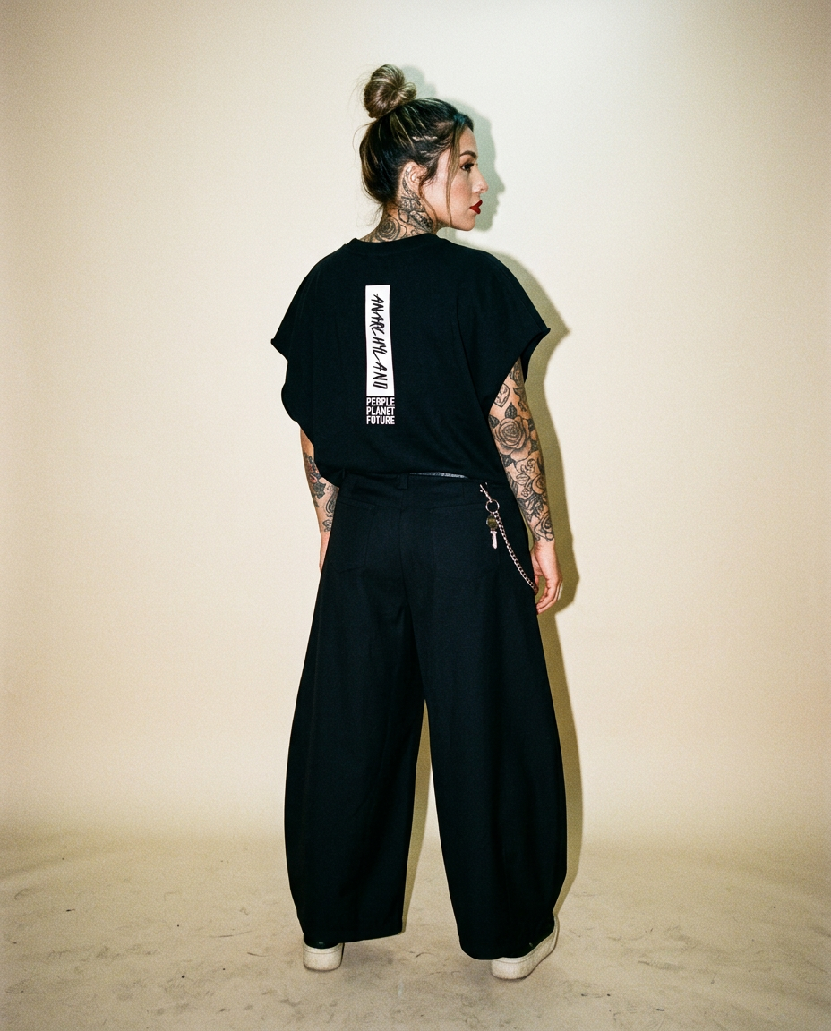 Person with tattoos, back turned, wearing a black oversized T-shirt and wide-leg pants in a studio; vertical white text on the back reads 'PEOPLE PLANET FUTURE'