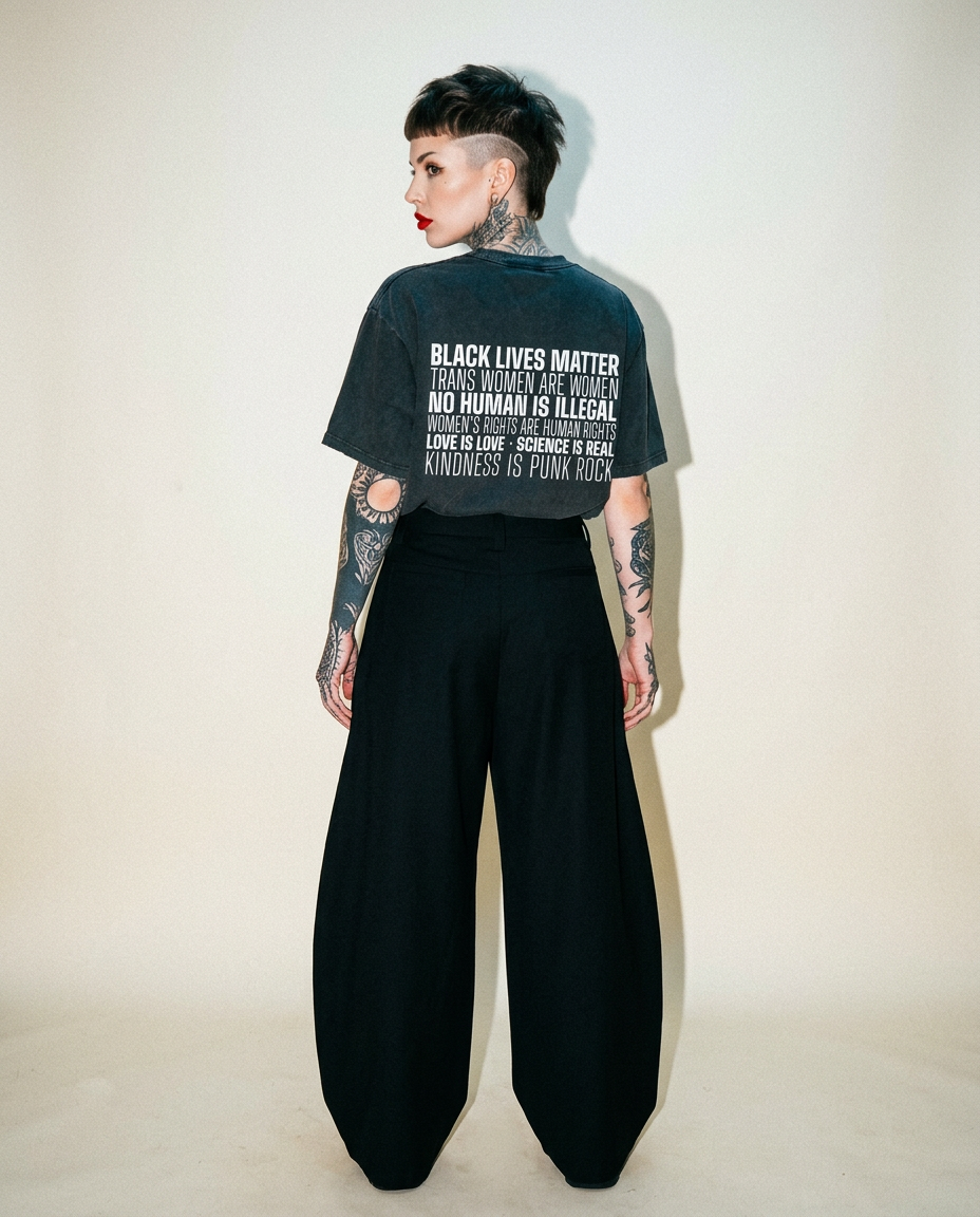 Back view of a person with a cropped dark T-shirt featuring bold white social-justice text, black wide-leg pants, and visible tattoos on arms and neck.