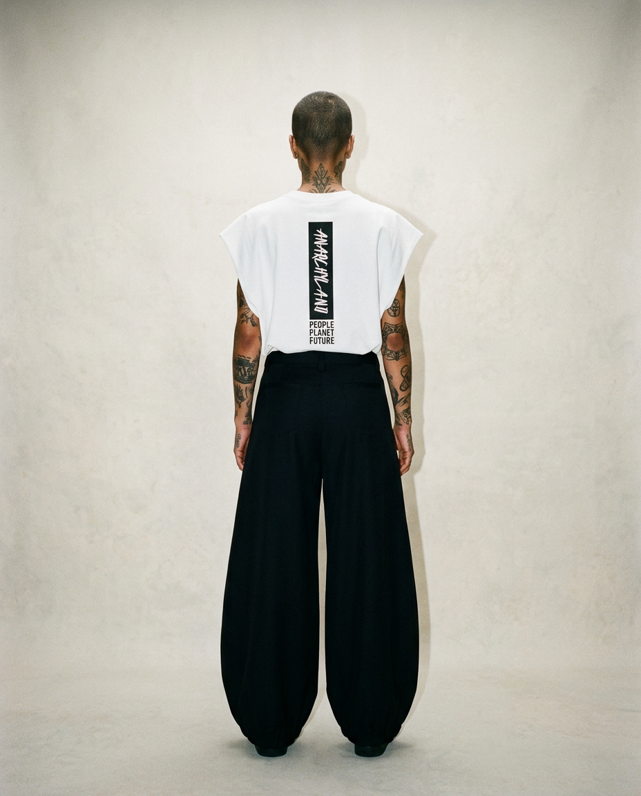 Person seen from behind in a white sleeveless top with a vertical black logo on the back and wide black balloon pants; tattoos on arms.