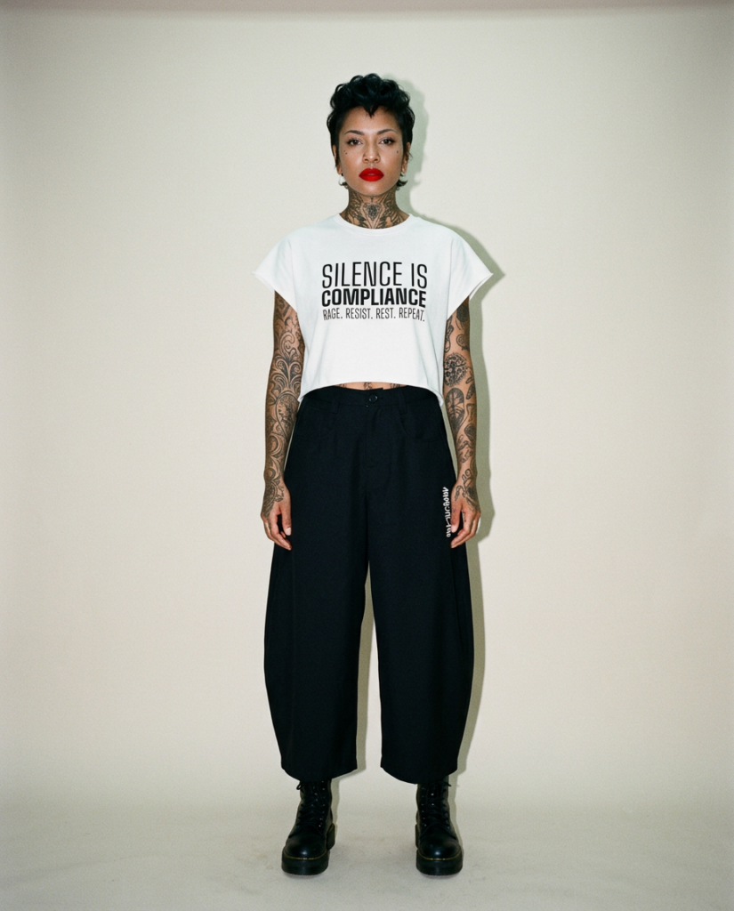 Model with short dark hair and red lipstick faces the camera wearing a white cropped t shirt that reads silence is compliance with black wide leg pants and combat boots showing full tattoos on arms and neck in a studio setting