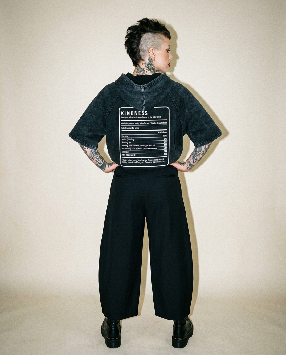 Person with a mohawk and visible tattoos, back view wearing a black oversized jacket with a 'KINDNESS' graphic on the back panel.