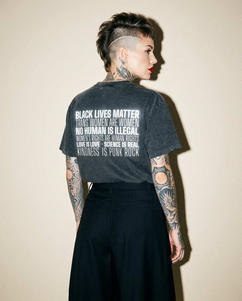 Person with a punk mohawk and tattoos, viewed from behind, wearing a dark T-shirt with bold social-justice text on the back.
