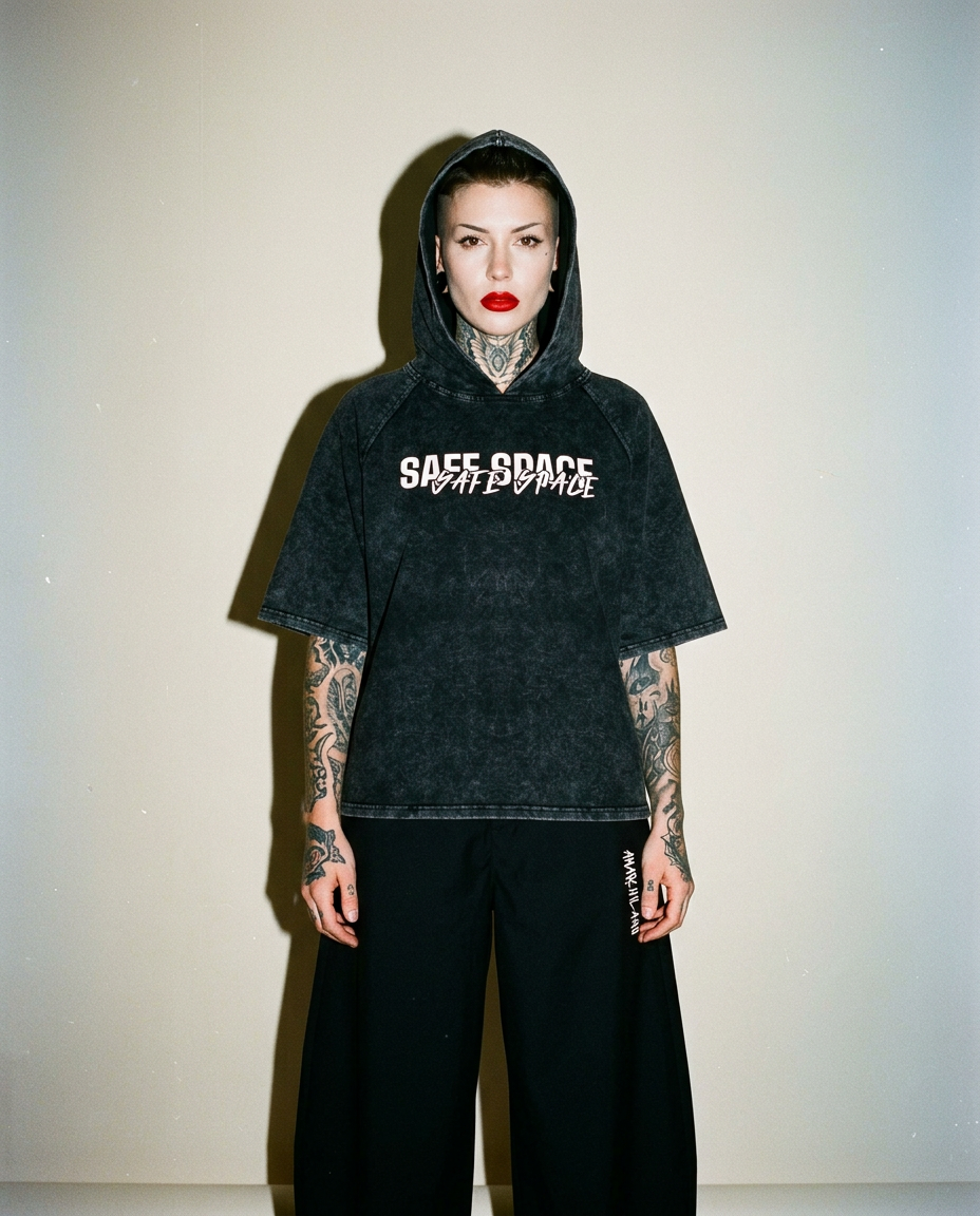 Person wearing a dark, oversized hoodie with a white graphic, black wide-leg shorts, bold red lipstick, and full-arm tattoos standing against a pale wall.