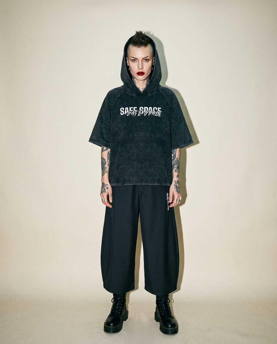 Person stands facing the camera in an oversized charcoal hoodie with 'SAFE SPACE' text, black baggy pants, combat boots, and tattooed arms.