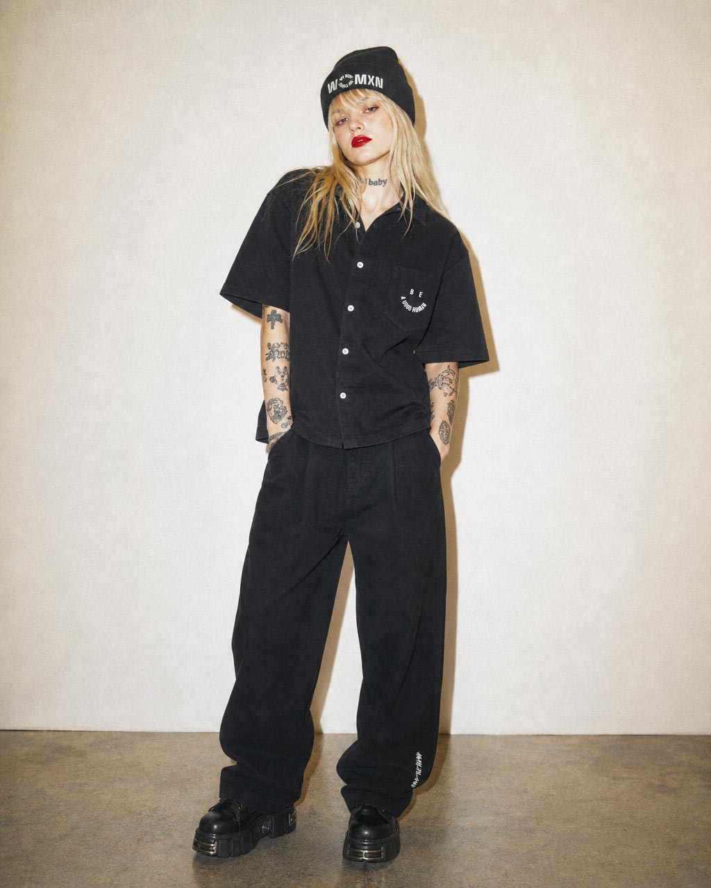 Person in all black streetwear pose against a beige wall black button up shirt baggy black pants chunky black shoes and a black beanie with white text visible tattoos on arms and neck