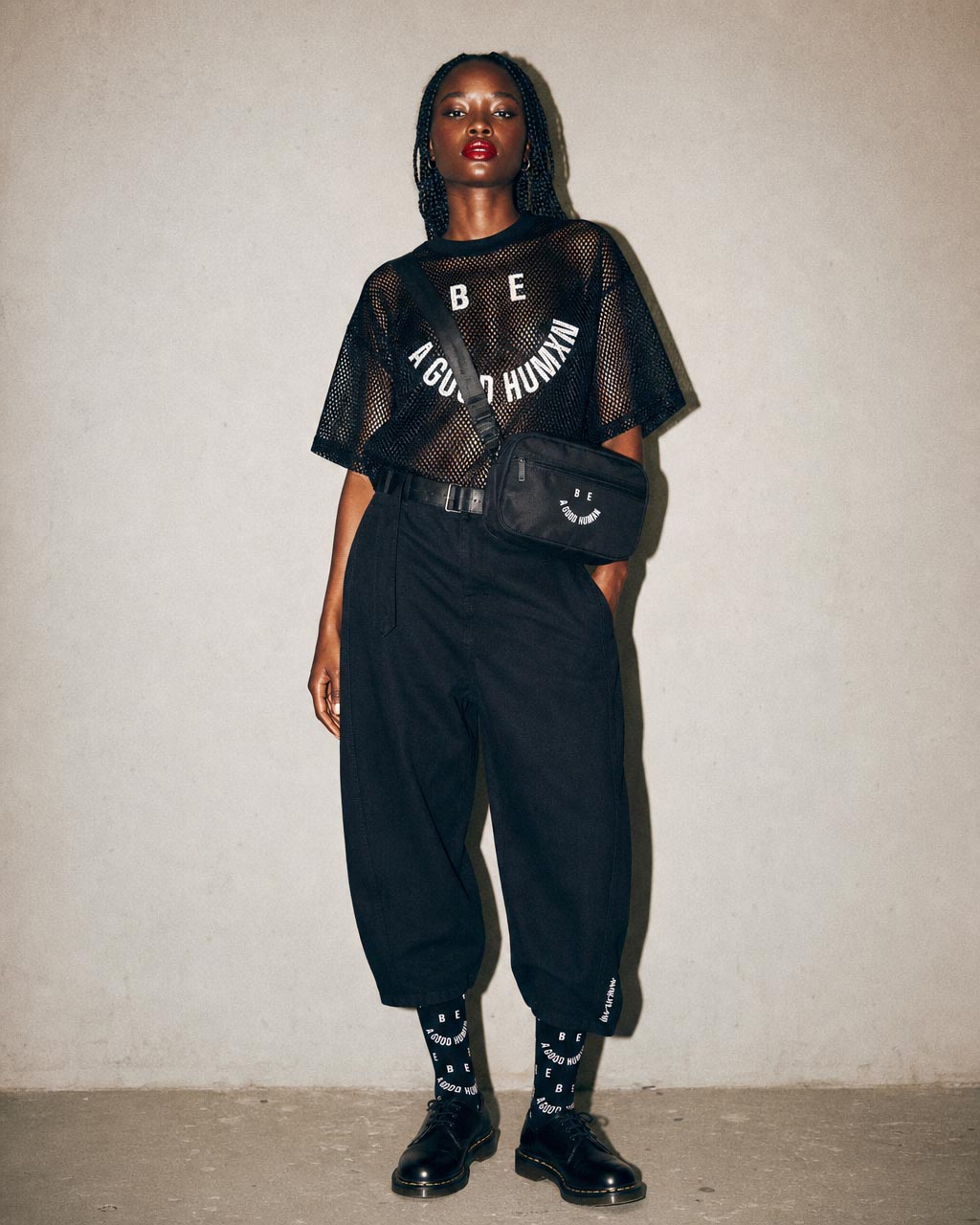 Woman in a black mesh tee with be a good human text high waisted black pants crossbody bag and black boots posing against a neutral wall
