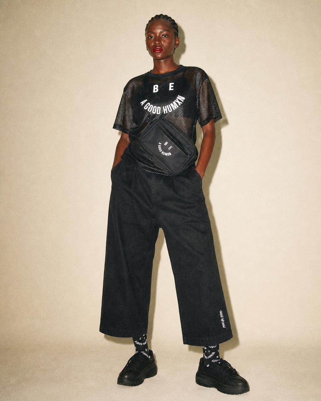 Person standing against a beige backdrop wearing a black mesh t shirt that reads be a good human black wide leg pants sneakers and a small crossbody bag worn diagonally across the chestwaist