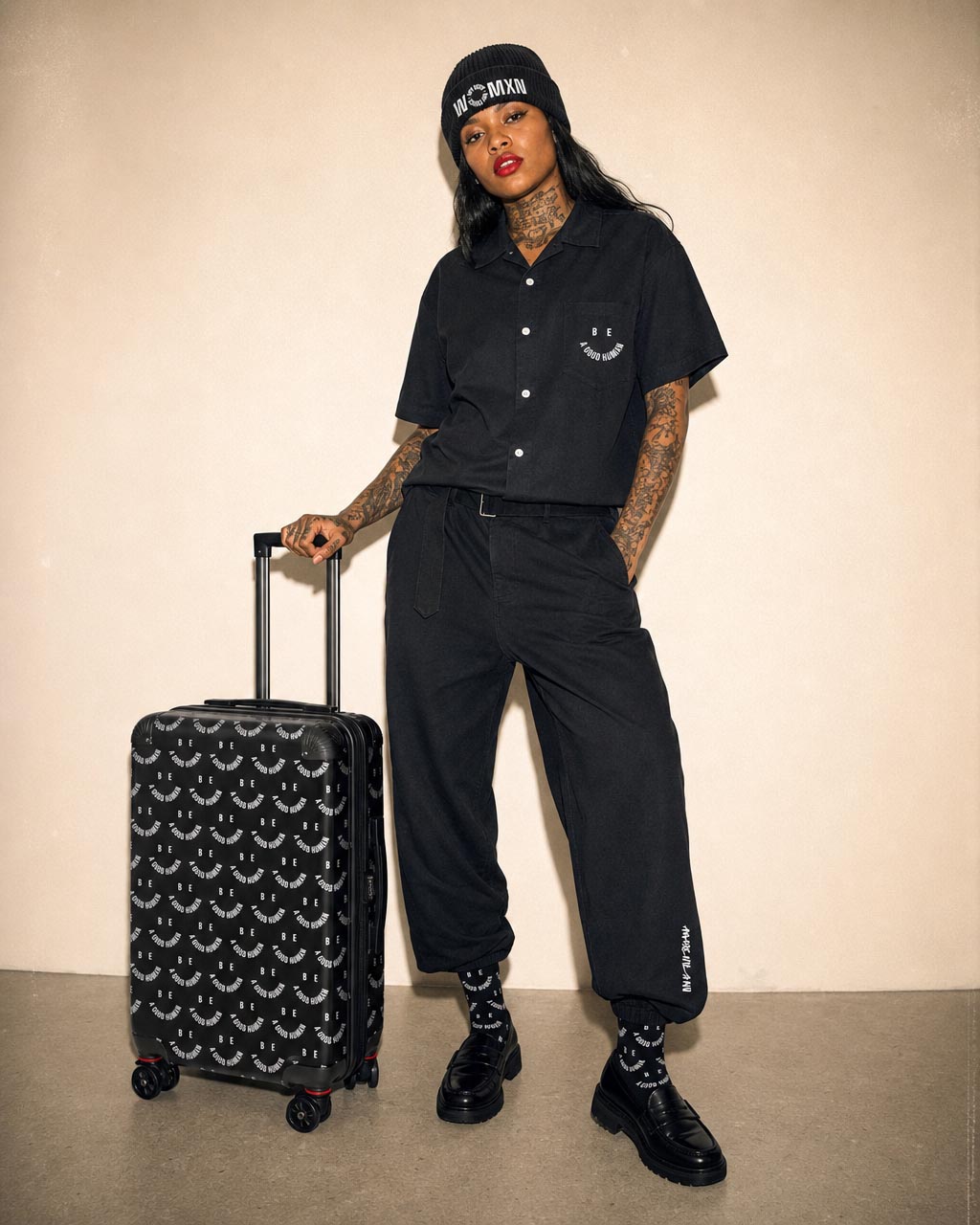 Model posing in all black travel outfit with a patterned rolling suitcase wearing a beanie and cuffed pants showing tattoos on arms and neck