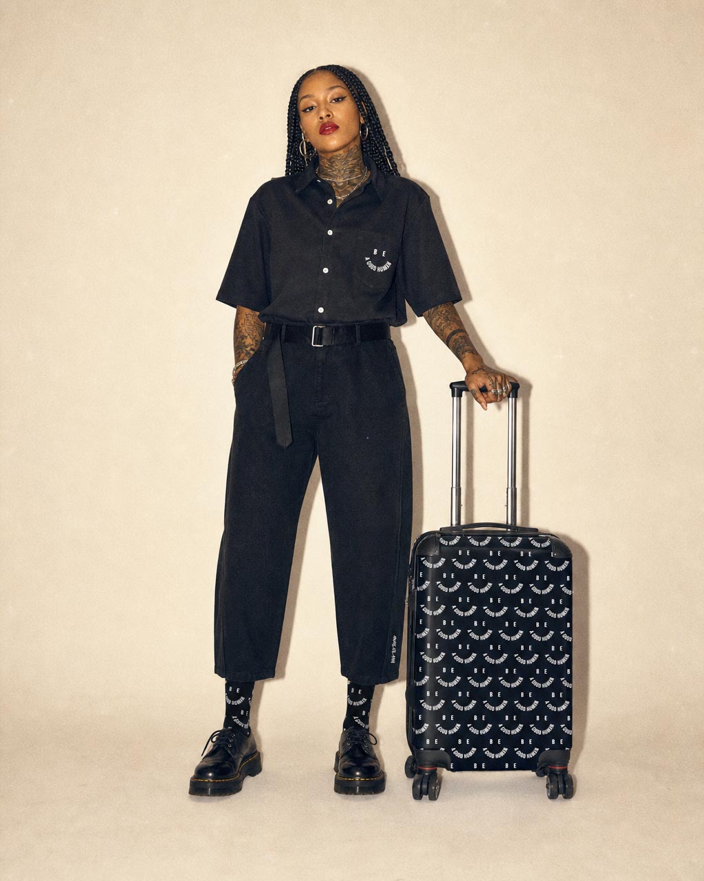Person in an all black outfit with braids and visible tattoos stands with a patterned rolling suitcase against a plain beige background