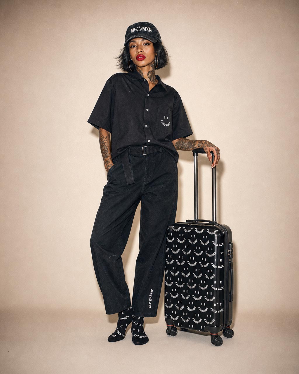 Woman in an all black outfit with a cap and bold red lipstick posing with a black patterned rolling suitcase beside her