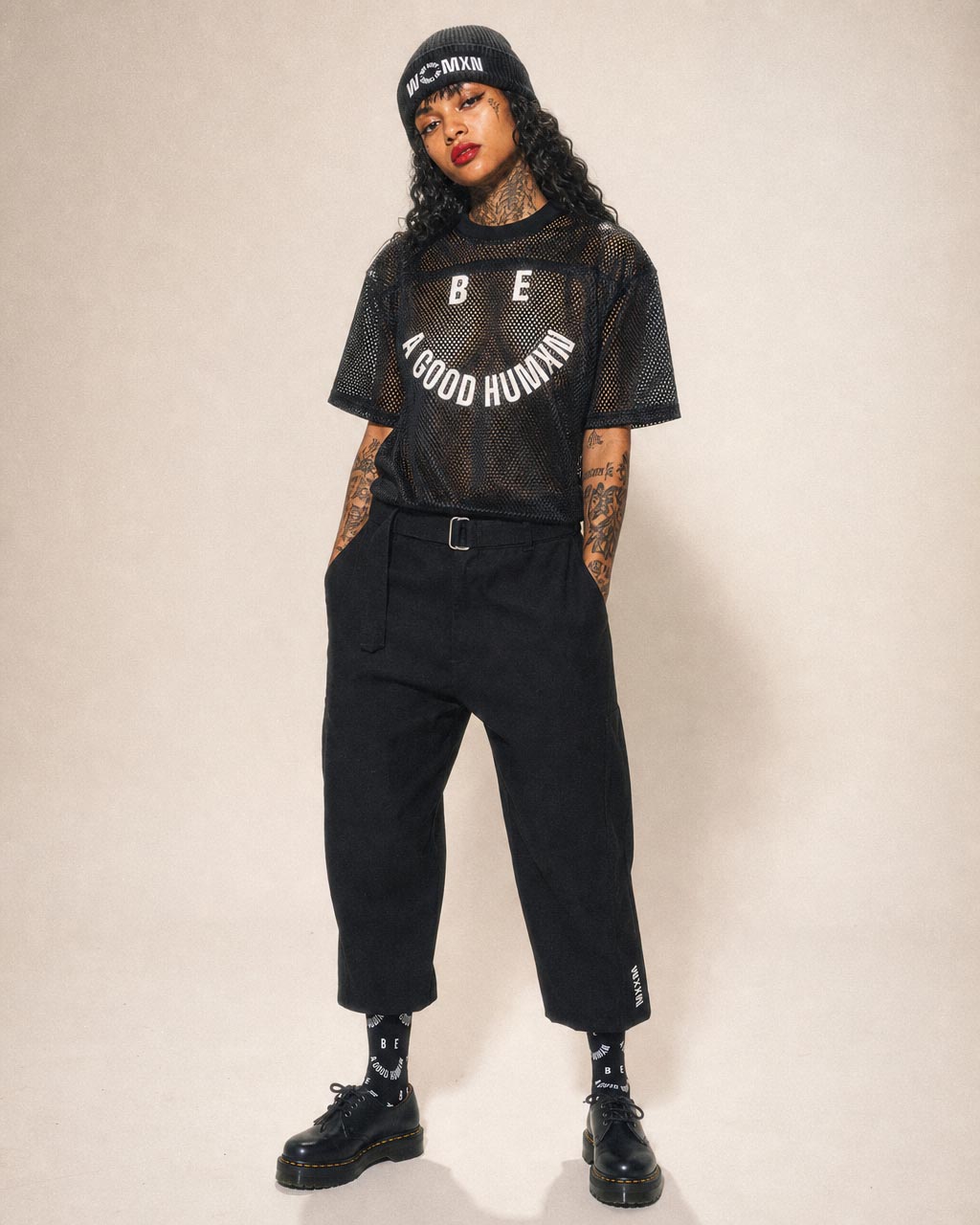 Person wearing a black mesh tshirt with the slogan be a good human black belt pants beanie and boots posing against a neutral background