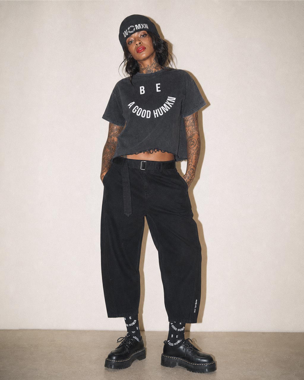 Person standing against a beige wall wearing a cropped black tshirt that reads be a good human in a circular layout black pants chunky black shoes a black beanie and visible tattoos on arms