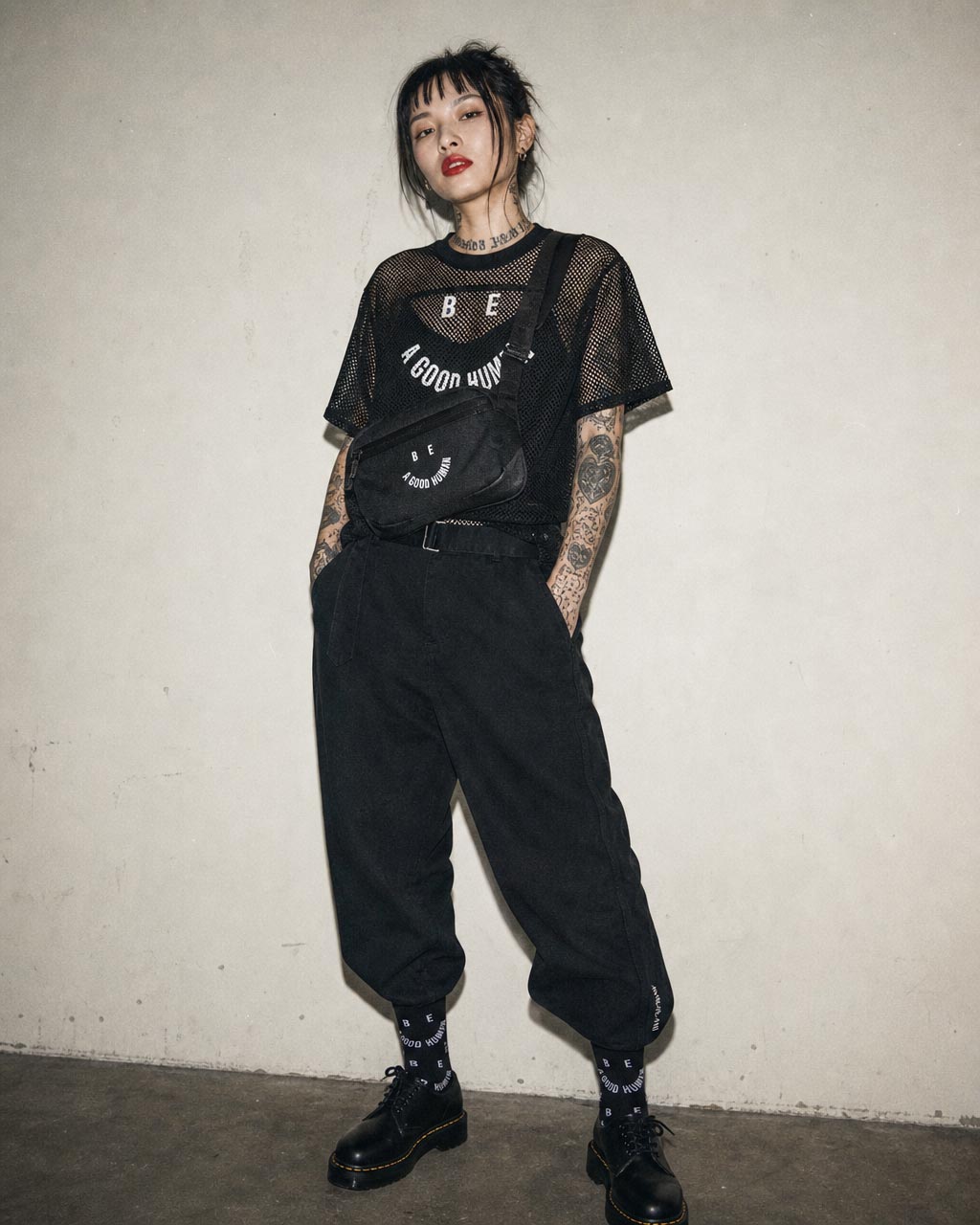 Young person in an all black streetwear outfit mesh top baggy pants and a crossbody fanny pack standing against a plain wall with hands in pockets and visible arm tattoos