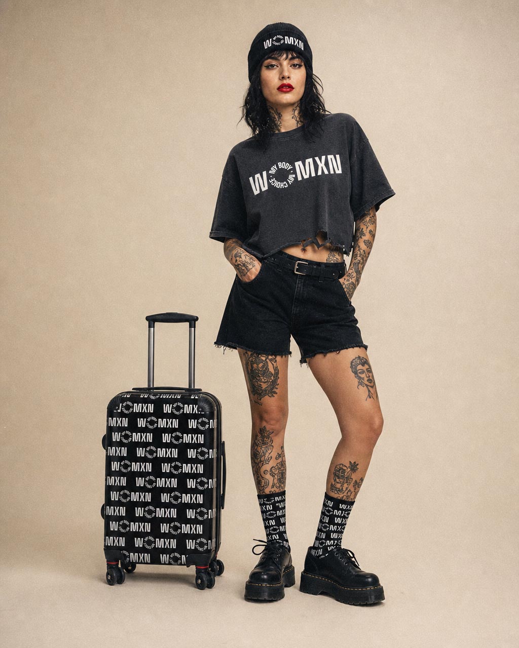 Tattooed woman in a black cropped tee shorts beanie and chunky boots posing with a patterned rolling suitcase against a beige backdrop