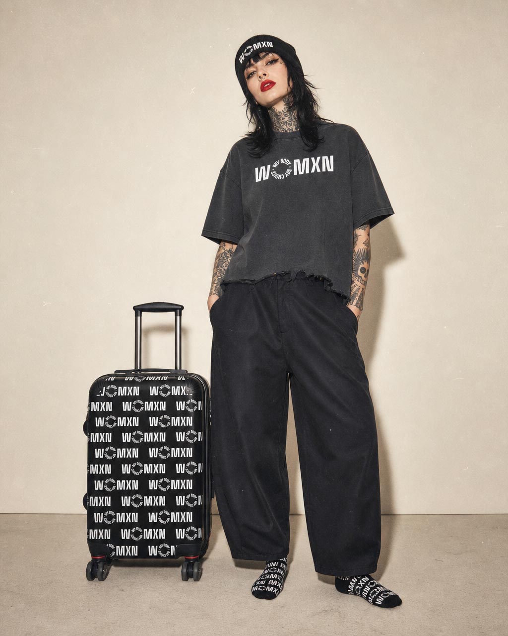 Model in a black oversized tee and wide black pants tattoos visible standing with a black rolling suitcase covered in white logos