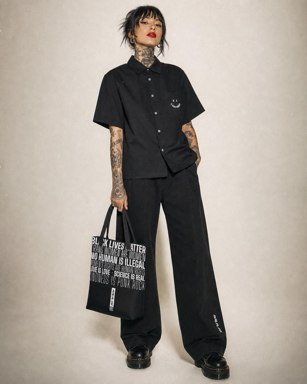 Person wearing a loose black shirt and wide leg black pants holding a black tote bag with white text standing against a neutral backdrop