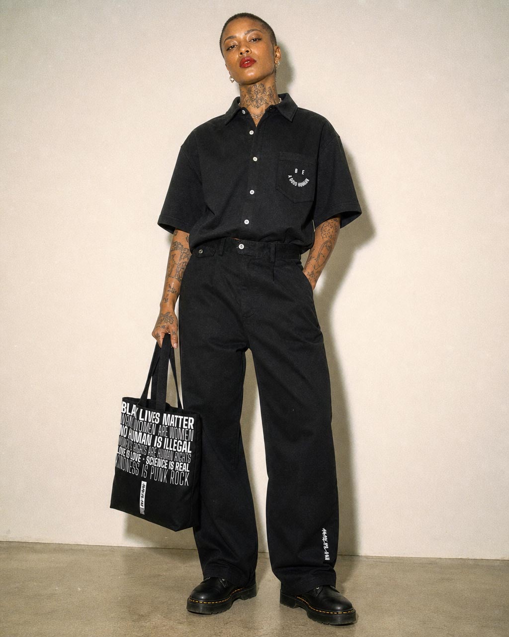 Model wearing all black streetwear oversized short sleeve button up shirt with logo pocket wide leg pants black shoes and a black tote reading black lives matter
