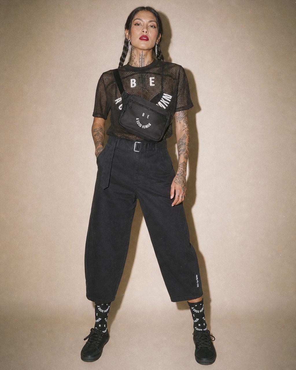 Model wearing a black mesh top wide black pants and a crossbody bag with bold lettering against a beige backdrop posing confidently