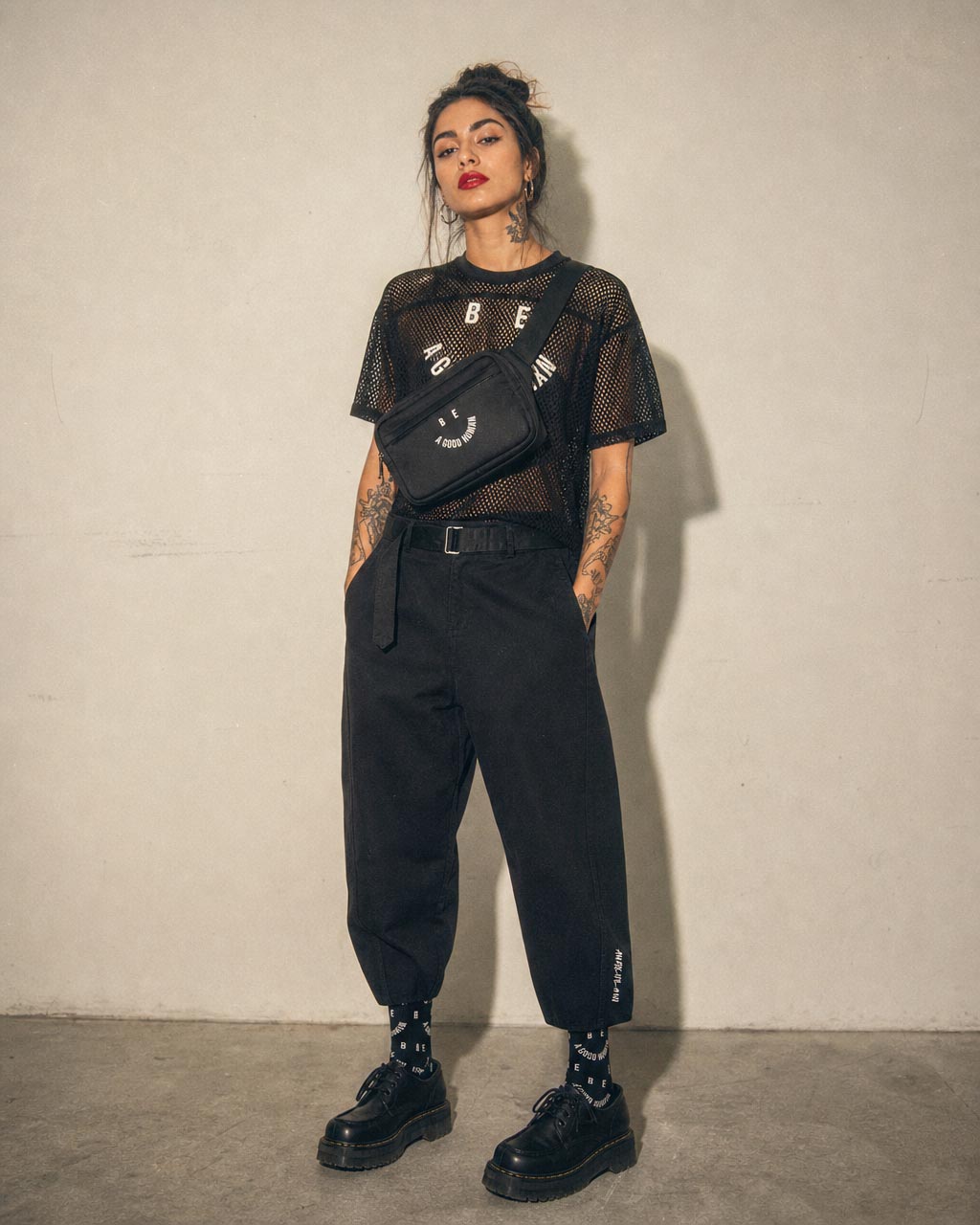 Woman with dark hair in a mesh black top and belted black pants wearing a small crossbody bag and chunky shoes posed against a beige wall