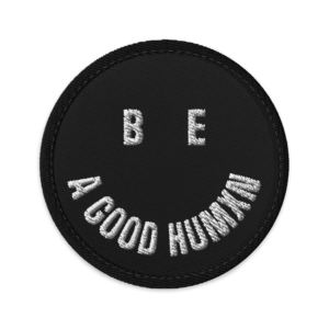 Close-up of a black circular fabric badge with white embroidered letters along the edge, on a black background.