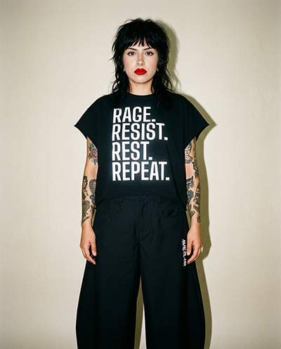 Person with black hair and tattoos wearing a black T-shirt that reads 'RAGE. RESIST. REST. REPEAT.' against a light wall.